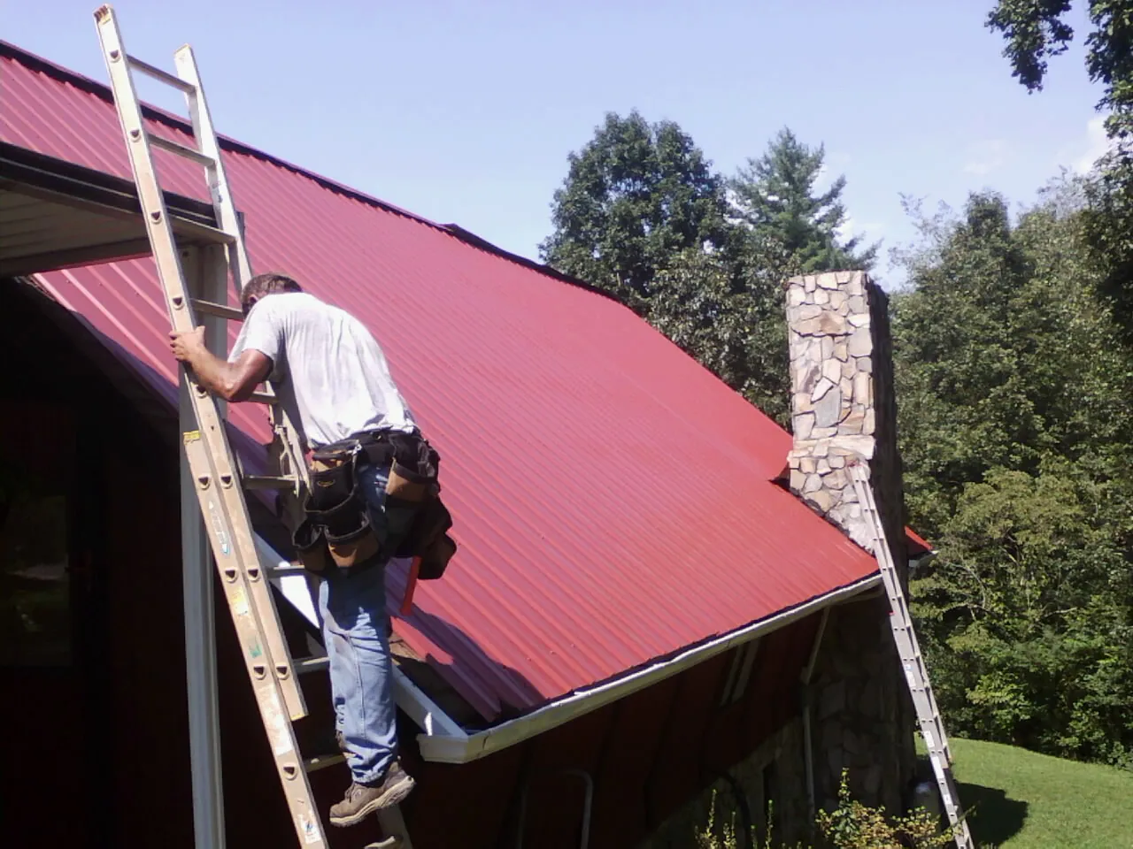 Roofer on a ladder beside a red metal roof and stone chimney — Soffit & Fascia Repair by Roofing Brothers in Cascadia